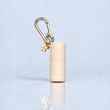 A beige cylindrical cosmetic case with a gold carabiner clip and a small white flower charm with yellow accents, standing on a reflective light blue surface.